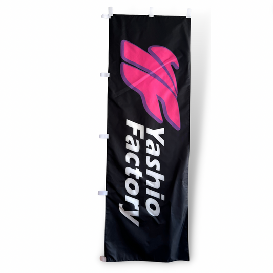 A rectangular flag with a black background featuring a pink graphic and the text 'YASHIO FACTORY' in white.